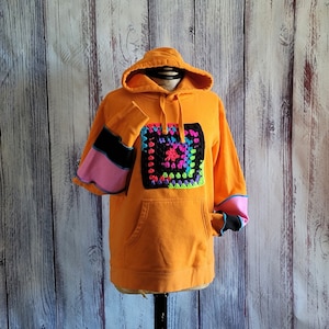 May include: Orange hoodie with a large, colorful crochet square on the front. The sleeves have pink, black, and blue accents. The hoodie has a front pocket and a hood with a drawstring.