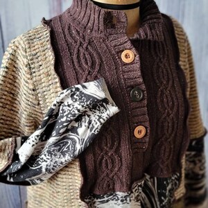 May include: A patchwork cardigan with a high collar and wooden buttons. The sweater features a mix of textures and patterns, including cable knit, woven fabric, and a black and white print. The color palette includes shades of brown, beige, and black.