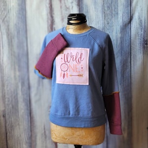 May include: A blue sweatshirt with maroon colored sleeves. The sweatshirt has a pink patch with the text "Wild One" and a dreamcatcher design.
