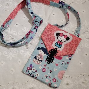 May include: A small, rectangular crossbody bag with a flap closure, featuring a geisha design. The bag is made of light blue fabric with a floral pattern and a pink flap. The strap is a combination of pink and blue fabrics.