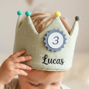 May include: A light green fabric crown with a polka dot pattern, featuring a blue and white number 3 emblem. The crown has a name embroidered in black script and is topped with colorful pom-poms. A child is wearing the crown.