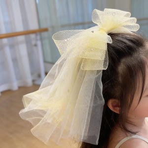 Puede incluir: Un lazo para el pelo de color amarillo claro con un diseño en capas y un velo transparente. El lazo está adornado con pequeños adornos brillantes. El accesorio está diseñado para ser usado en el cabello.