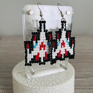 May include: A pair of pixelated earrings featuring a white, red, black, and blue design. The earrings are made of small beads and hang from silver-colored hooks. The earrings are displayed on a clear acrylic stand.