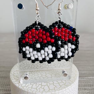 May include: A pair of beaded earrings in a Poké Ball design. The earrings feature a red, black, and white color scheme, with a silver hook. The earrings are displayed on a clear acrylic stand.