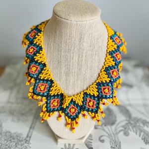 May include: A vibrant beaded necklace displayed on a mannequin. The necklace features a repeating geometric pattern in yellow, teal, red, and black beads. The necklace is a statement piece, perfect for adding a pop of color to any outfit.