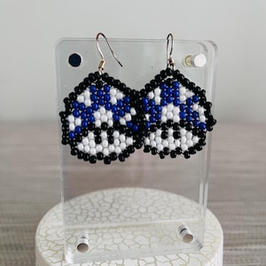 May include: Handmade beaded earrings with a geometric design. The earrings feature a pattern of blue, white, and black beads. The earrings are displayed on a clear acrylic stand, showcasing the intricate beadwork and craftsmanship.