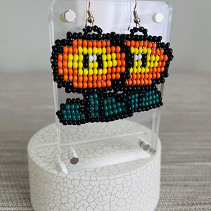 May include: A pair of beaded earrings featuring a pixelated flower design. The earrings have orange, yellow, black, and teal beads, with silver-colored ear wires. The earrings are displayed on a clear acrylic stand.