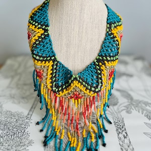 May include: A vibrant beaded necklace displayed on a mannequin. The necklace features a turquoise base with yellow, red, and black accents. Long beaded fringe in turquoise, yellow, orange, and red hangs from the bottom, creating a bold statement piece.