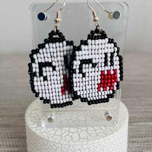 May include: A pair of handmade pixel art earrings featuring a ghost design. The earrings are made with white, black, and red beads, and have gold-colored fish hook ear wires. The earrings are displayed on a clear acrylic stand.