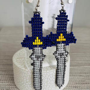 May include: A pair of beaded earrings shaped like swords. The earrings feature a blue hilt with yellow accents and a silver blade. The earrings are displayed on a clear stand.