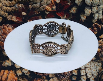 May include: A gold-toned metal bracelet with an intricate, openwork design. The bracelet is on a white surface.
