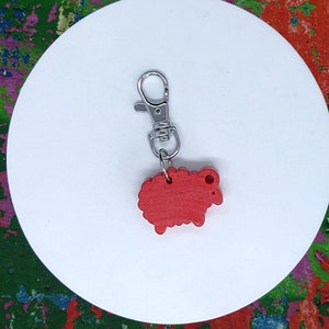 Könnte beinhalten: Ein roter schaförmiger Anhänger mit einem silberfarbenen Verschluss. Der Anhänger ist aus Holz gefertigt und hat ein einfaches, cartoonartiges Design. Der Verschluss ist am Kopf des Schafs befestigt. Der Anhänger liegt auf einer weißen, kreisförmigen Oberfläche.