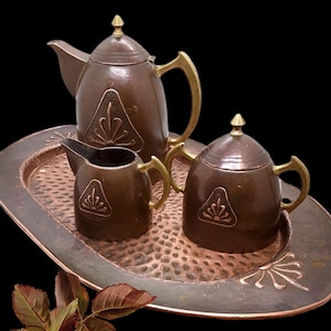 May include: A copper tea set comprising a teapot, a creamer, and a lidded sugar bowl, each featuring brass handles and finials. The items are arranged on an oval, hammered copper tray with a decorative fan motif, alongside a small copper leaf sprig.