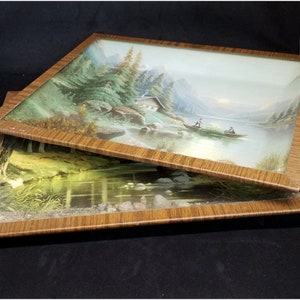 Op de afbeelding: Twee ingelijste landschapschilderijen met houten lijsten. Het bovenste schilderij toont een scène met een meer met bergen op de achtergrond en twee mensen in een boot. Het onderste schilderij toont een bos scène met een meer en bomen.