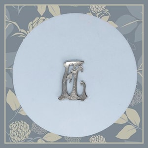 Puede incluir: Un amuleto de metal de color plateado en forma de la letra "E". La letra es adornada y tiene un diseño detallado.