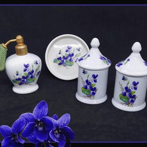 Peut inclure: Un ensemble de vanité en porcelaine blanche comprenant un flacon de parfum, deux pots à couvercle et une petite soucoupe. Chaque pièce est ornée d'un motif floral de violettes violettes et de feuilles vertes. Le flacon de parfum a un vaporisateur doré et un couvercle en tissu vert.
