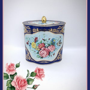 Könnte beinhalten: Eine blaue und goldene Dose mit einem Blumendesign. Die Dose hat einen gewellten Rand und einen goldenen Deckel mit einem Knopf. Die Blumen sind rosa, gelb und weiß.