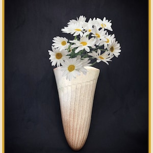 May include: A white ceramic vase with a textured surface holds a bouquet of white daisies. The vase is shaped like a cone and has a narrow opening. The daisies are arranged in a loose, natural style.