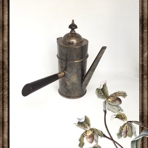 Op de afbeelding: Een zilveren koffiekan met een houten handvat en een decoratieve bovenkant. De kan staat op een wit oppervlak met een boeket groene en bruine orchideeën.