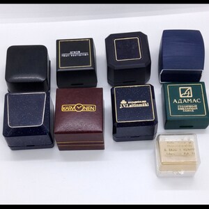 Op de afbeelding: Een assortiment van negen kleine, vierkante, rechthoekige en achthoekige ringdozen in verschillende kleuren, waaronder zwart, blauw, groen, bruin en goud. De dozen hebben verschillende merknamen erop gedrukt, waaronder "Stella", "Karvonen", "J.V. Laitinmaki" en "Adamas".