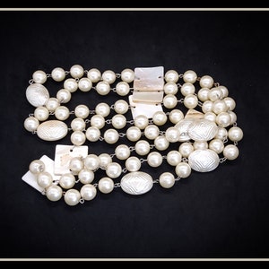 Puede incluir: Un collar de perlas de múltiples hilos con detalles de concha de forma ovalada. El collar está hecho de perlas blancas y tiene un diseño de concha de color crema.
