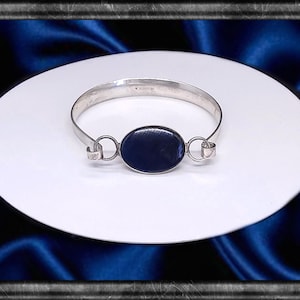 Könnte beinhalten: Silberner Armreif mit einem dunkelblauen, ovalen Stein in der Mitte. Der Armreif wird auf einer weißen Oberfläche vor einem dunkelblauen Hintergrund präsentiert. Das Design ist schlicht und elegant.
