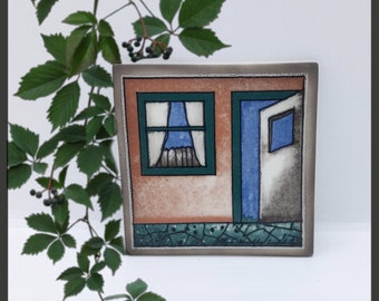 May include: A ceramic tile with a painted image of a small house with a blue door and a window with a blue curtain. The house is painted on a peach background with a green trim.