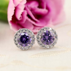 May include: A pair of round stud earrings with a central purple gemstone surrounded by a halo of smaller, clear stones. The earrings are set in a silver-colored metal and rest on a white surface, with pink flowers in the background.