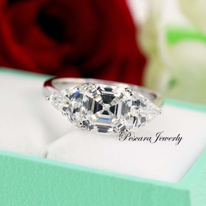Puede incluir: Un anillo de plata con un gran diamante de forma cuadrada y dos diamantes triangulares más pequeños a cada lado. El anillo está en una caja azul claro con el texto "Pescara Jewelry" en la esquina inferior derecha.