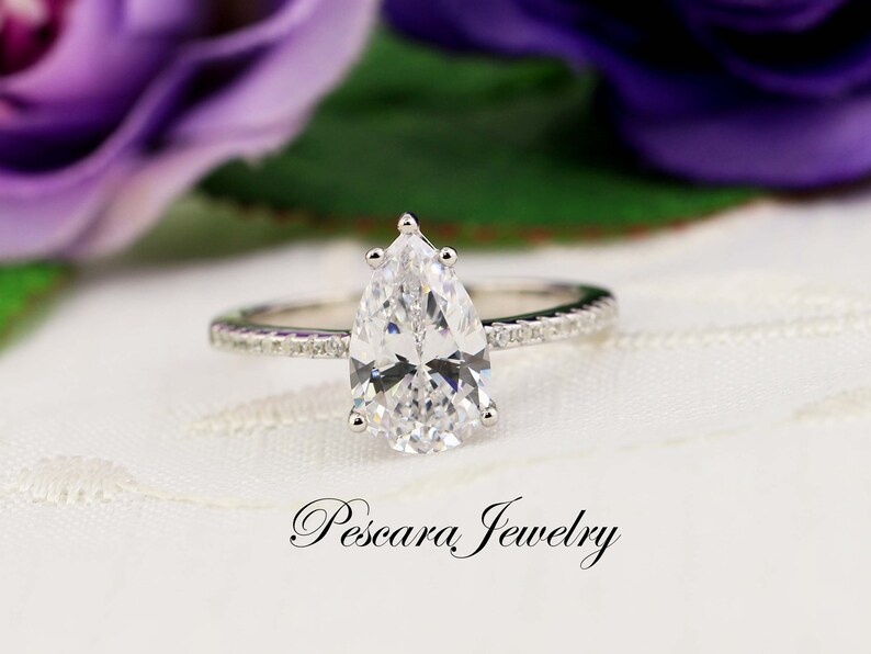 May include: A pear-shaped diamond engagement ring with a pave band. The ring is set in silver and features a large, clear diamond.