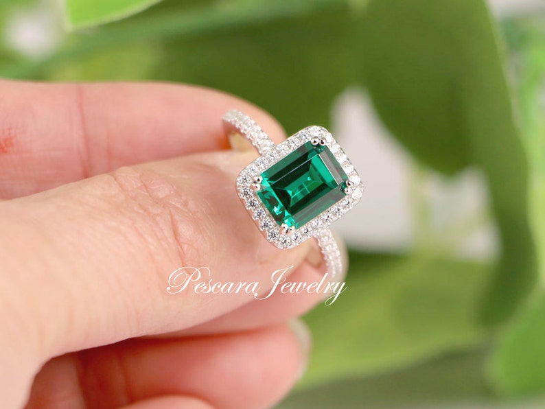 May include: A silver ring with a large emerald green gemstone surrounded by smaller clear gemstones. The ring is set in a halo style.