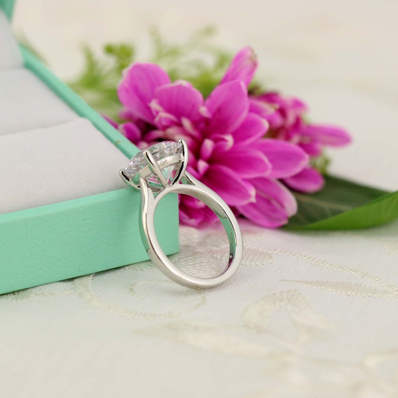 May include: A silver ring with a large, clear, round gemstone set in a prong setting. The ring is sitting on a white surface with a pink flower behind it.