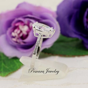 May include: A silver ring with a pear-shaped clear stone and smaller clear stones set in the band. The ring is on a white display stand with the text "Pescara Jewelry" on it.