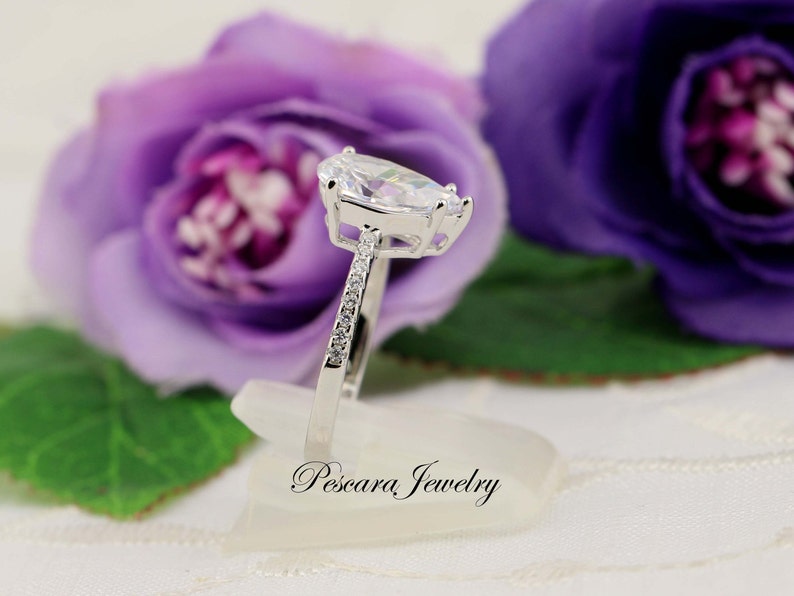May include: A silver ring with a pear-shaped clear stone and smaller clear stones set in the band. The ring is on a white display stand with the text "Pescara Jewelry" on it.
