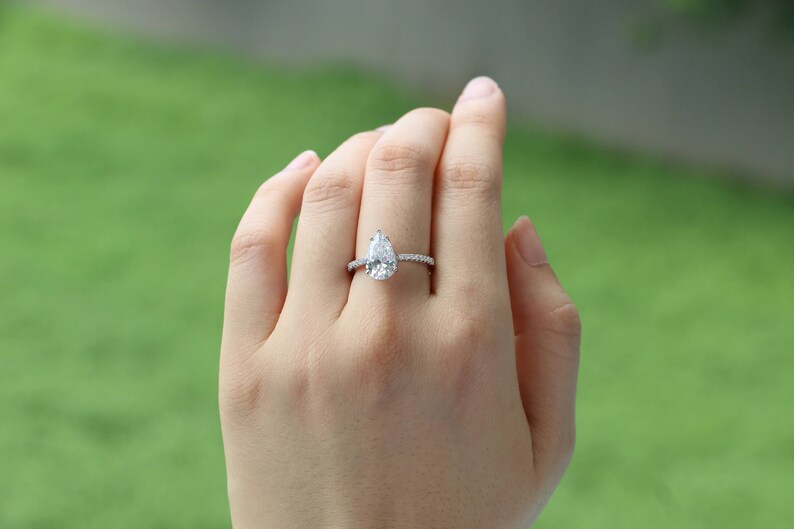 May include: A pear-shaped diamond engagement ring with a pave setting. The ring is set in a silver band.