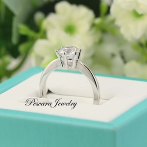 May include: A silver ring with a single, round, clear gemstone in a prong setting. The ring is in a turquoise box with the words "Pescara Jewelry" printed on the inside.
