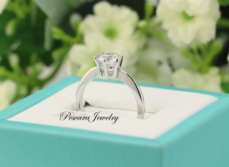 May include: A silver ring with a single, round, clear gemstone in a prong setting. The ring is in a turquoise box with the words "Pescara Jewelry" printed on the inside.