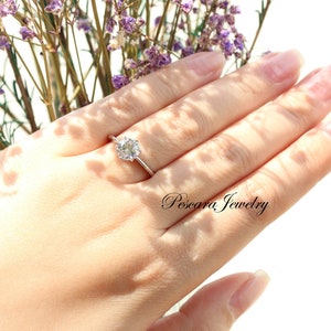 May include: A silver ring with a round, clear gemstone on a finger. The ring is set against a background of purple flowers.