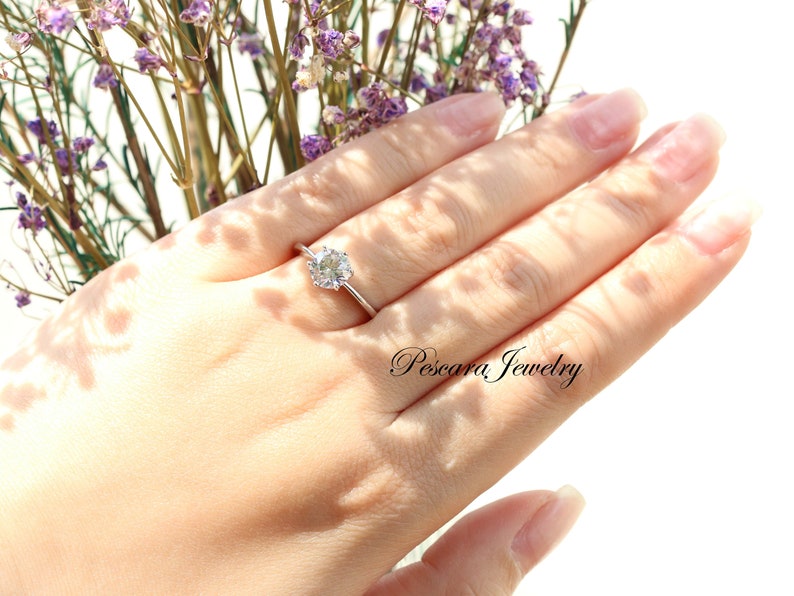 May include: A silver ring with a round, clear gemstone on a finger. The ring is set against a background of purple flowers.