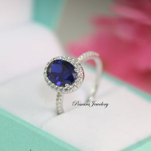 May include: A silver ring with a large oval-shaped blue sapphire surrounded by a halo of smaller white diamonds. The ring is set on a white background.