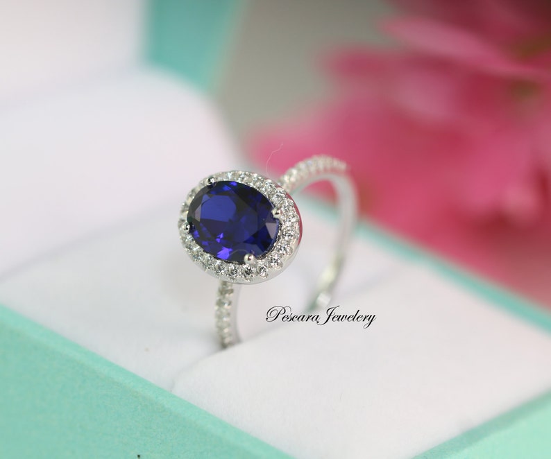 May include: A silver ring with a large oval-shaped blue sapphire surrounded by a halo of smaller white diamonds. The ring is set on a white background.
