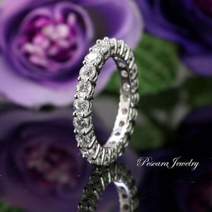 Puede incluir: Un anillo de eternidad plateado con una fila continua de piedras preciosas redondas y claras. El anillo se encuentra sobre un fondo borroso de flores moradas y una superficie reflectante. Joyería de Pescara Jewelry.