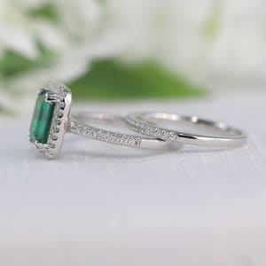 May include: A silver engagement ring set with a large emerald green gemstone surrounded by smaller clear gemstones. The ring is paired with a matching band with smaller clear gemstones.