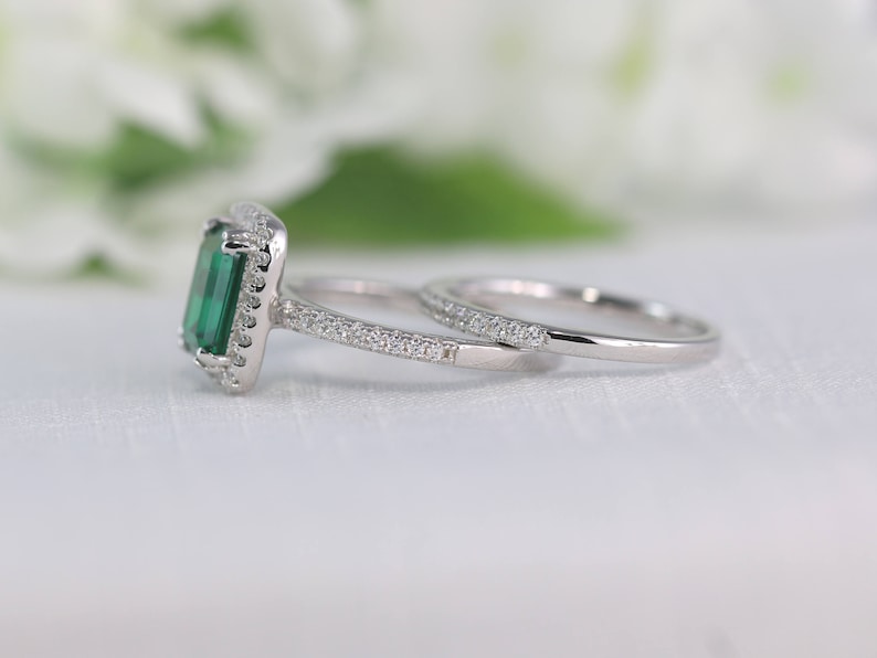 May include: A silver engagement ring set with a large emerald green gemstone surrounded by smaller clear gemstones. The ring is paired with a matching band with smaller clear gemstones.