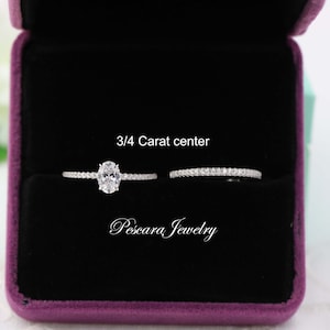 Peut inclure: Une bague de fiançailles en argent avec un diamant central ovale de 3/4 carat et une bande assortie avec de petites pierres rondes. Les bagues sont présentées dans un écrin à bijoux en velours violet. Le texte "3/4 Carat center" et "Pescara Jewelry" est visible sur l'image.