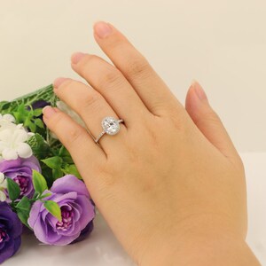 Op de afbeelding: Een zilveren ring met een ovale middelste steen omringd door kleinere stenen. De ring zit aan een vinger.