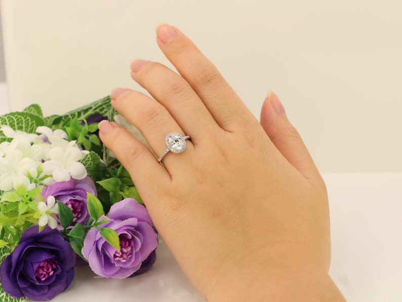 Op de afbeelding: Een zilveren ring met een ovale middelste steen omringd door kleinere stenen. De ring zit aan een vinger.