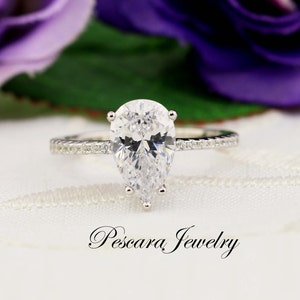 May include: A pear-shaped diamond engagement ring with a silver band and a row of smaller diamonds on the band. The ring is set on a white background with a purple flower in the background.