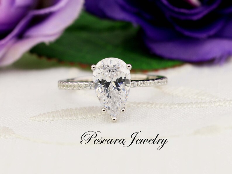 May include: A pear-shaped diamond engagement ring with a silver band and a row of smaller diamonds on the band. The ring is set on a white background with a purple flower in the background.