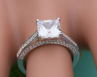 Puede incluir: Un anillo de compromiso de plata con un gran diamante de forma cuadrada y una doble fila de diamantes más pequeños alrededor de la banda.